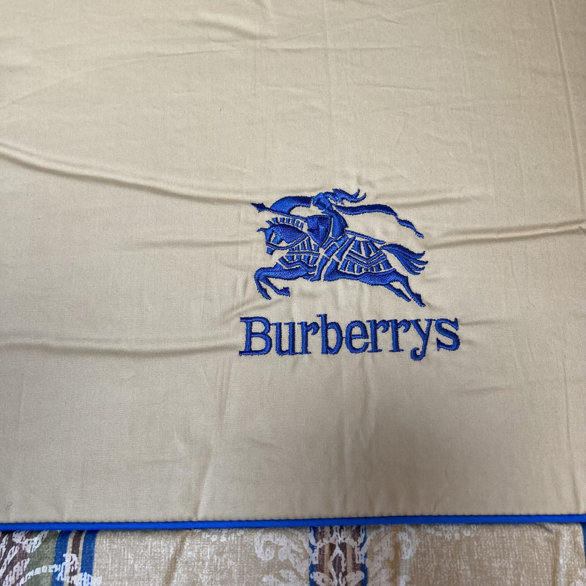 Royal blue embroidered Burberrys Equestrian Knight logo and lettering on beige fabric.