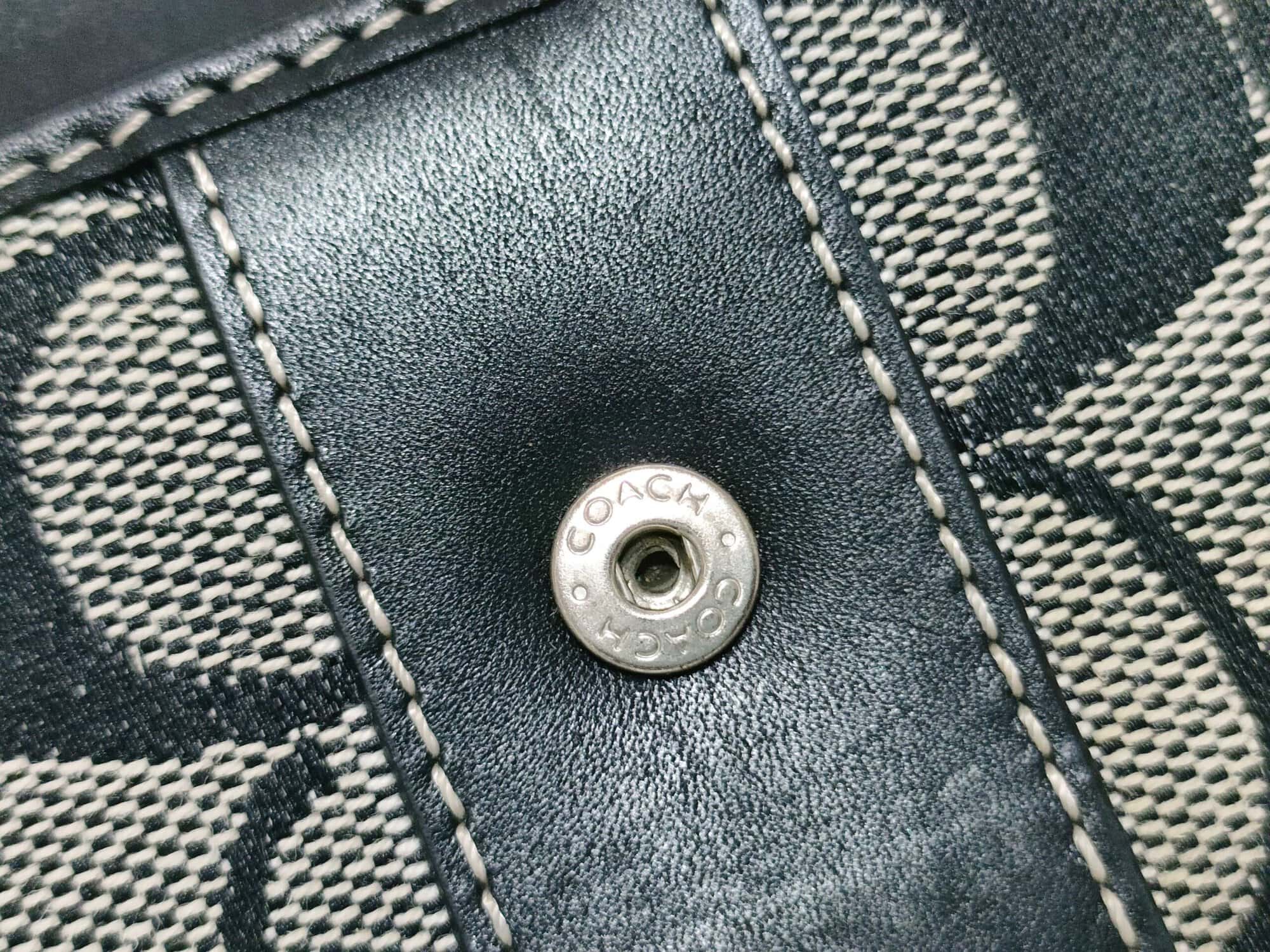 Close-up of engraved silver-tone Coach snap button on black leather stripe.